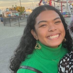 Smiling woman with long dark hair, wearing green turtleneck in front of a Ferris wheel.