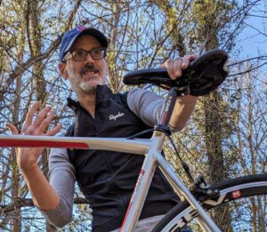 White man with glasses and ball cap holding bicycle.